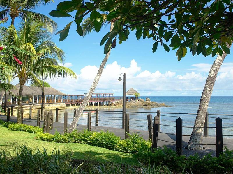 Strand mit Palmen, Holzzaun und blaugem Himmel, ruhiges Meer und grüne Wiese im Vordergrund.