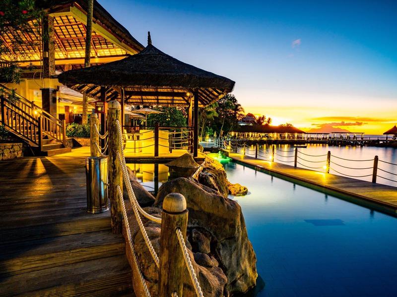 Ein beleuchtetes Holzdeck mit Pavillon neben einem ruhigen Pool und Sonnenuntergang am Wasser.