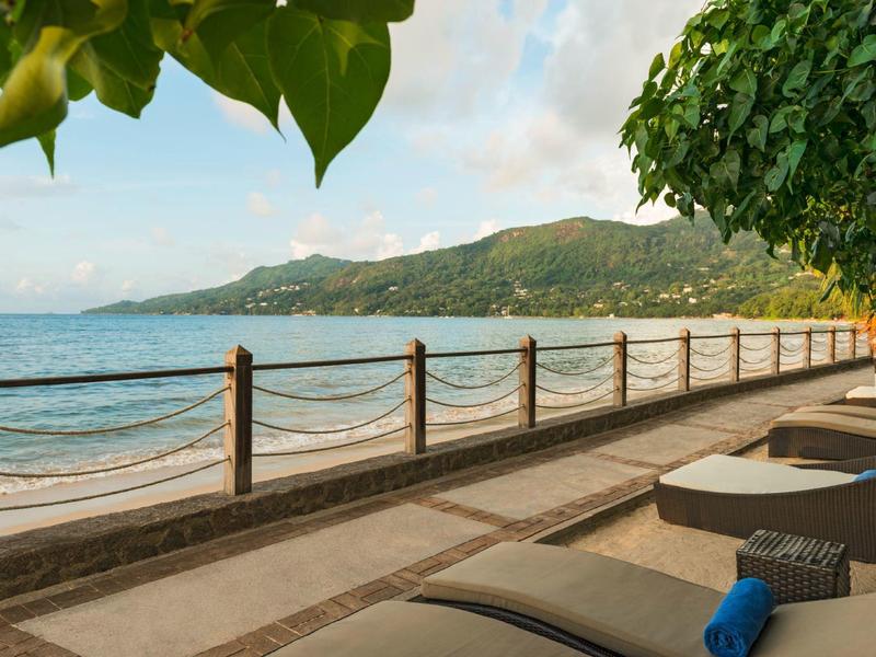 Küste mit Meerblick, Berglandschaft, Promenade und Liegestühlen unter grünen Blättern.