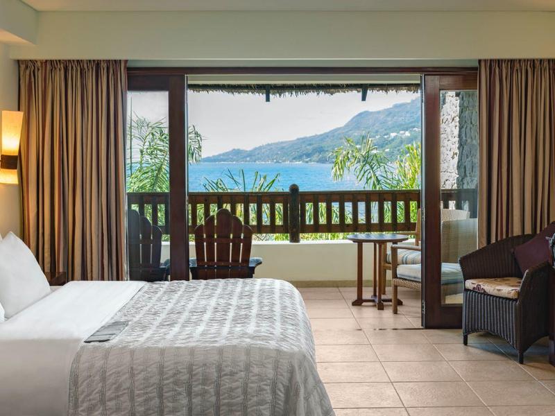 Hell eingerichtetes Schlafzimmer mit großem Bett, Balkon und Blick auf grüne Hügel und Berge.