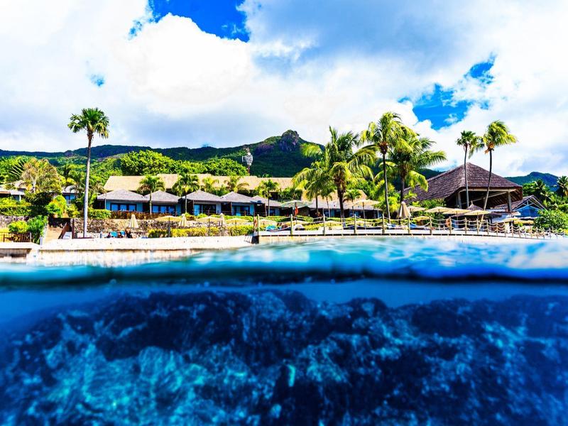 Resort am Strand mit Palmen, klarem Wasser und Korallenriff unter blauem Himmel und Wolken.