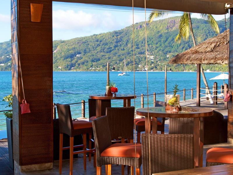 Offene Strandbar mit Holzstühlen und Tischen, Blick auf das blaue Meer und grüne Hügel im Hintergrund.