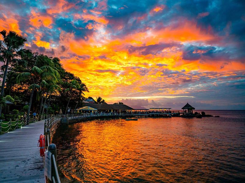 Sonnenuntergang über ruhigem Meer mit buntem Himmel, Palmen und einem Steg am Ufer.