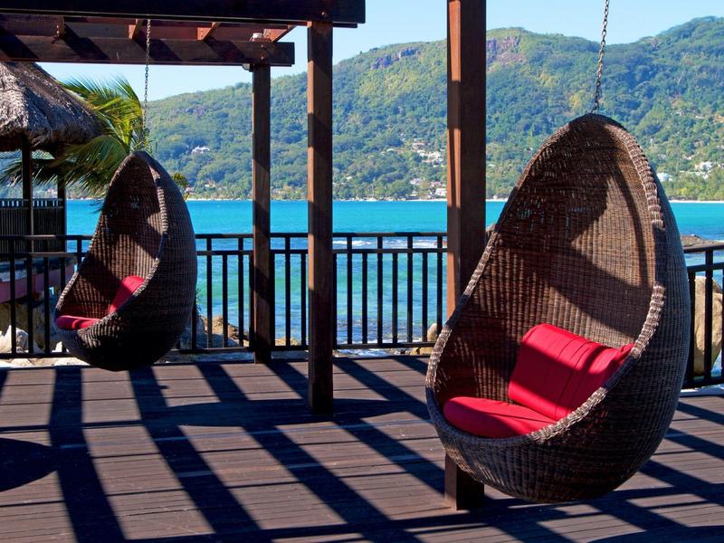 Zwei Rattanhängesessel mit roten Kissen auf Terrasse mit Blick auf blauen See und grüne Berge.