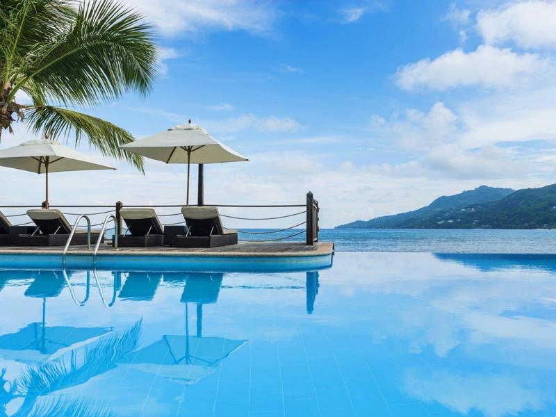 Ein Pool mit klarem Wasser, Sonnenschirmen, Palmen und Bergen im Hintergrund unter blauem Himmel.