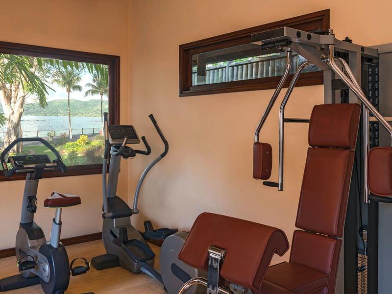 Kleiner Fitnessraum mit Trainingsgeräten, zwei Fenstern und Blick auf Palmen und Wasser draußen.