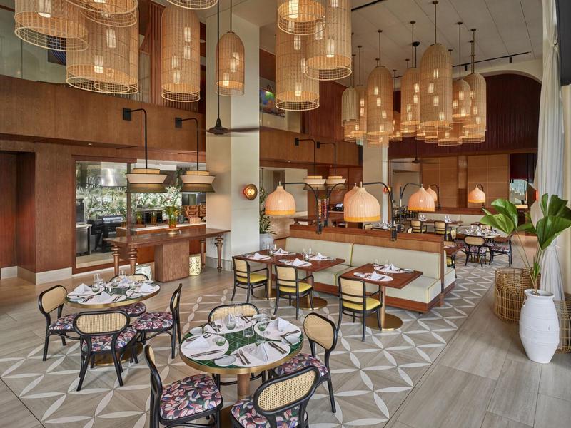 Modernes Restaurant mit runden Tischen, Holzstühlen, großen Lampen und offener Küche.