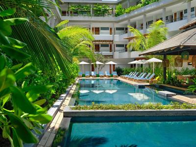 Hotelový bazén s lehátky a slunečníky obklopený zelení a budovami.