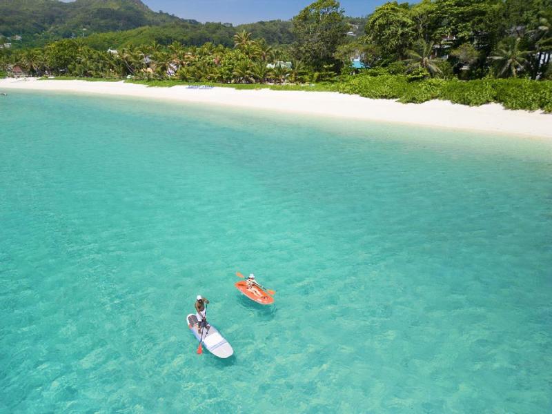 Čirá modrá laguna s dvěma pádlujícími na klidné vodě před bílou písečnou pláží a zelenými stromy.