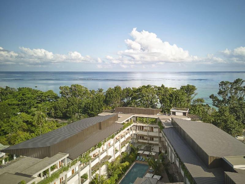 Výhled na hotelové letovisko s bazénem, obklopené stromy a mořem v pozadí.