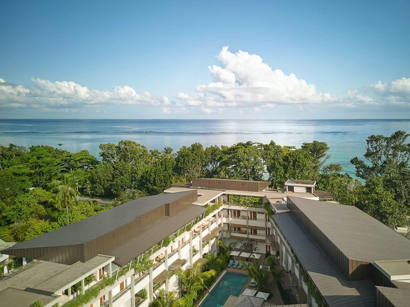 Pohled na resort s bazénem, obklopený stromy s výhledem na moře za jasného počasí.