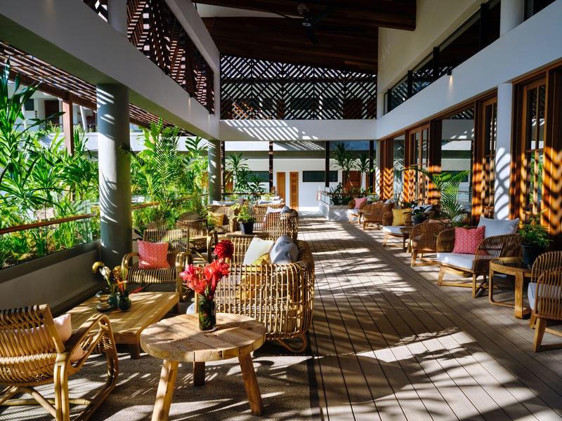 Světlý, otevřený hotelový salón s ratanovým nábytkem a rostlinami podél skleněné stěny.