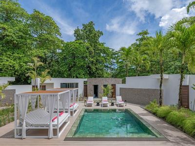 Modern pool area with loungers and a pavilion, surrounded by green trees and a white fence.