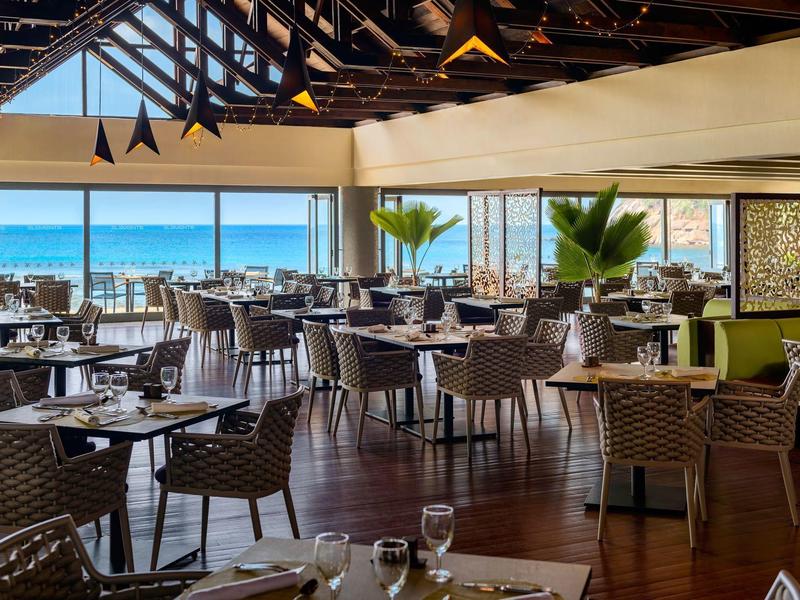 Modern restaurant with large windows, sea view, set tables, and wicker chairs.