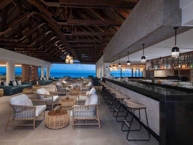 Modern hotel bar and lounge area with wooden roof and sea view at sunset.