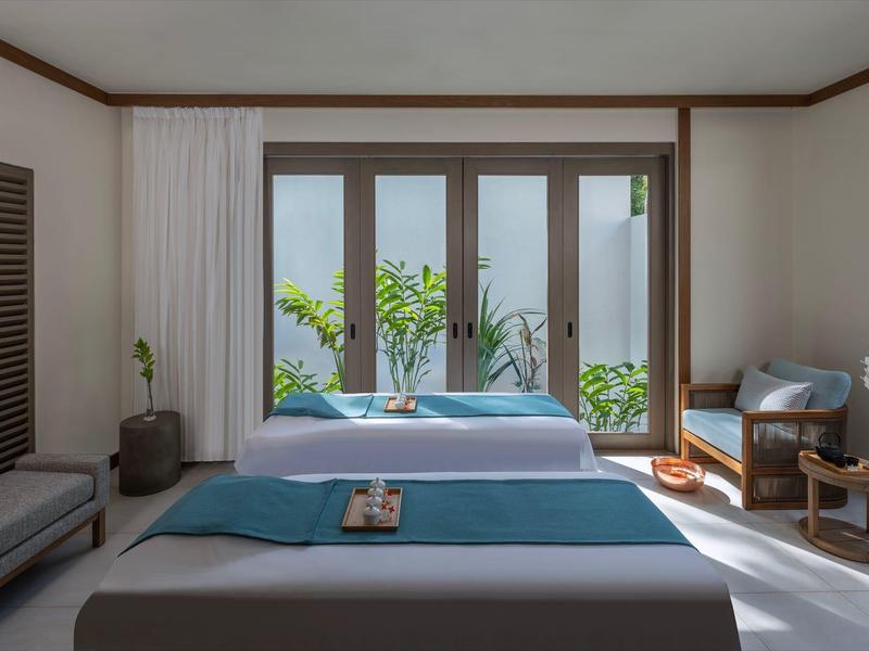 Bright hotel room with double bed, blue bedspread, and view of green plants through glass sliding doors.