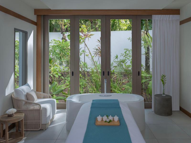 Bright wellness room with massage table, armchair, and large windows overlooking green plants.