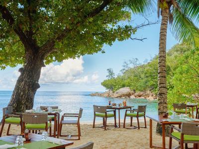 Restaurante en la playa con mesas y sillas de madera bajo palmeras junto al mar tranquilo y cielo azul.