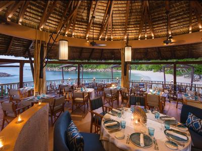 Restaurante abierto con techo de madera y vista al mar y playa de arena al atardecer.