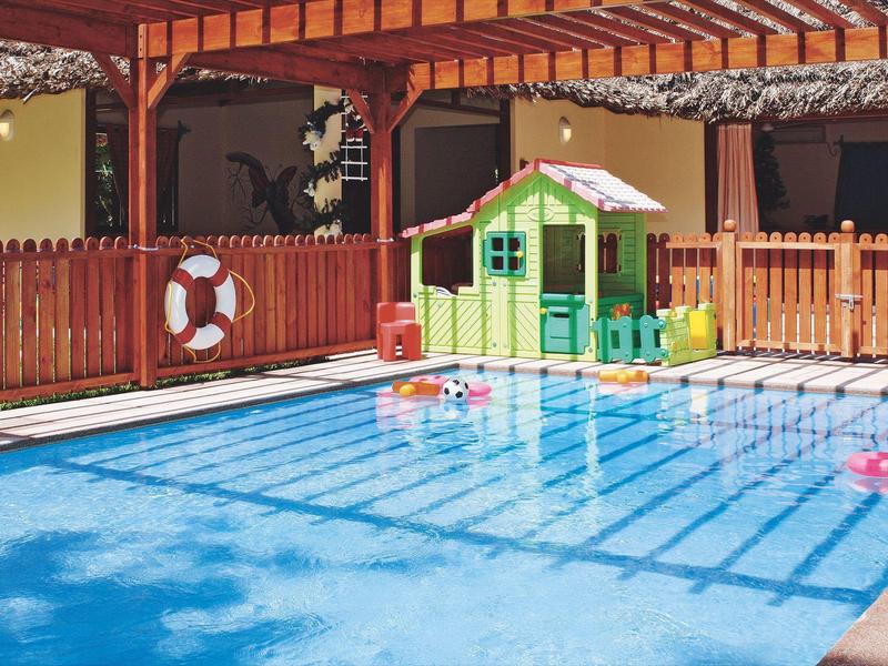 Área de juegos infantiles con piscina, casita de colores y zona soleada con sombra de vigas.