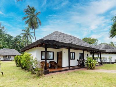 Ein Bungalow mit Strohdach und Veranda umgeben von Palmen und Grünfläche unter blauem Himmel.