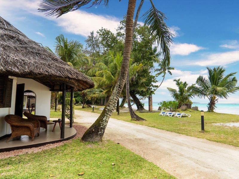 Resort sulla spiaggia con palme, prati, una strada lastricata e un edificio con tetto di paglia.
