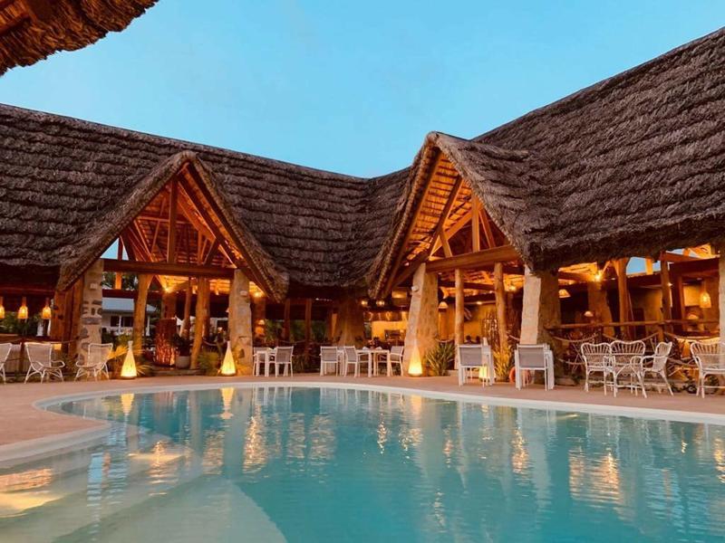 Restaurante del hotel iluminado con techo de paja y piscina al atardecer.