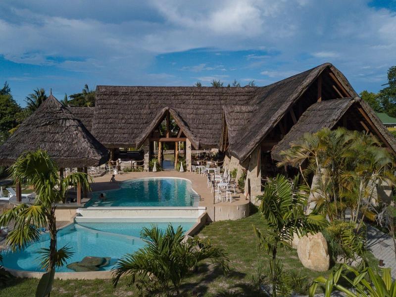 Lujosa cabaña con techo de paja, gran piscina y jardín tropical bajo cielo azul.