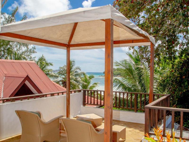 Terrasse mit drei weißen Stühlen, Holztisch, Pavillon und Blick auf Palmen und blauen Himmel.