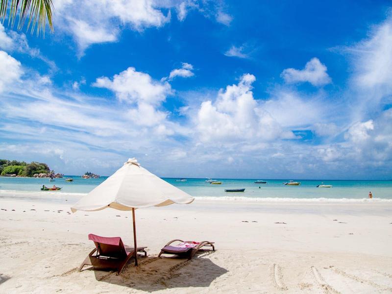 Strand mit weißem Sand, zwei Liegestühlen unter einem Sonnenschirm, blauem Himmel und ruhigem Meer.