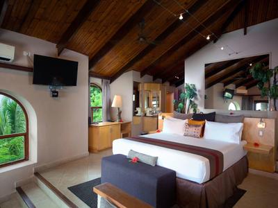 Chambre d'hôtel moderne avec plafond à poutres en bois, grand lit et vue sur des plantes vertes.