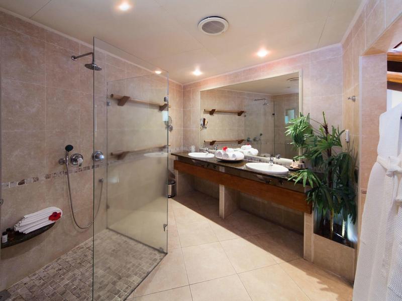 Salle de bain moderne avec douche en verre, doubles lavabos et grand miroir sous un éclairage chaud.