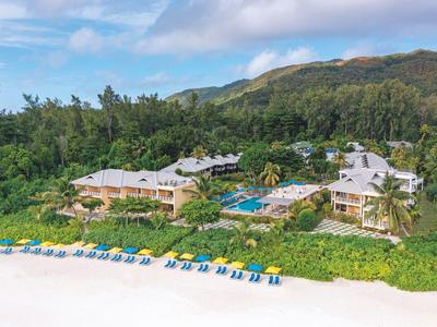 Resort mit mehreren Gebäuden, Pool, Palmen und gelben Strandliegen am weißen Sandstrand.