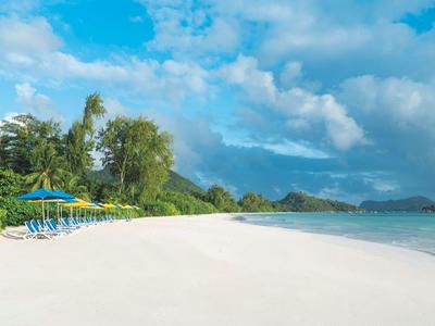 Breit wit zandstrand met blauwe parasols en palmbomen onder een blauwe lucht met wolken.