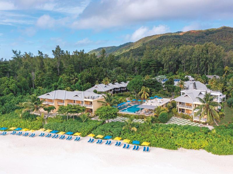 Hotelresort met zwembad aan het strand, omgeven door groen en bergen op de achtergrond.