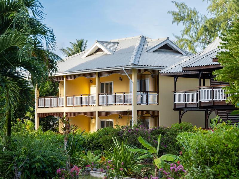 Geel huis met twee verdiepingen en veranda, omgeven door weelderige groene tuin.