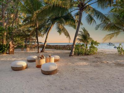 Strand mit Sand, Palmen und niedrigen Sitzkissen, im Hintergrund Meer und klarer Himmel.