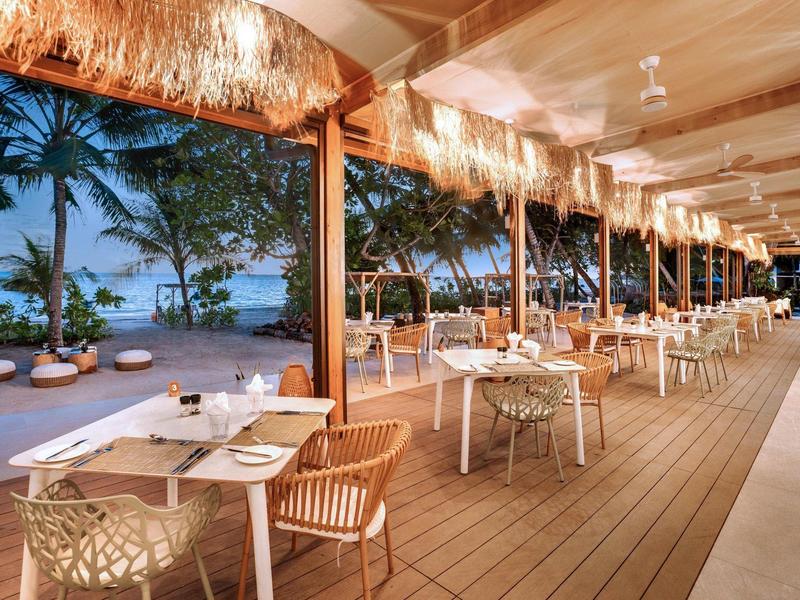 Restauracja na świeżym powietrzu z drewnianym tarasem i palmami przy plaży o zachodzie słońca.