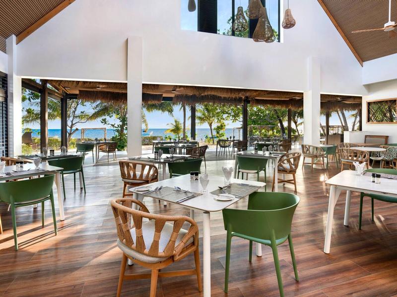 Jasna restauracja z drewnianą podłogą, zielonym i jasnym umeblowaniem oraz widokiem na morze.