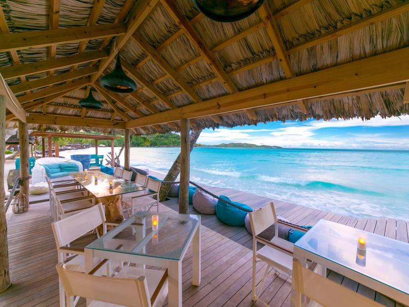 Restaurante de playa abierto con muebles de madera y vista al mar azul bajo un techo de paja.