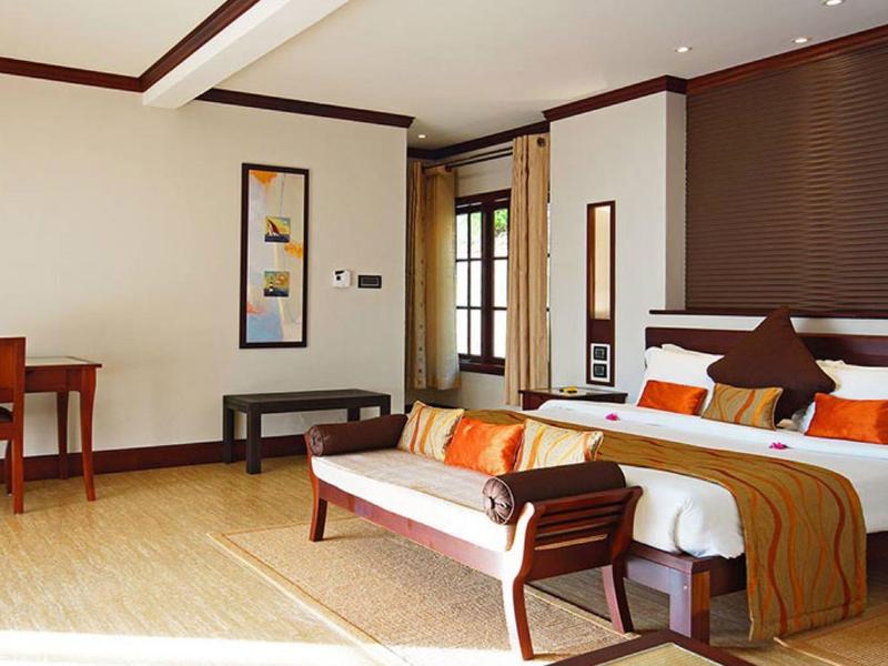 Habitación de hotel luminosa con cama grande, sofá y escritorio, madera clara y acentos cálidos.