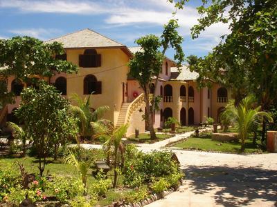Großes zweistöckiges Gebäude in beiger Farbe, umgeben von Grün und Pflasterweg bei blauem Himmel.