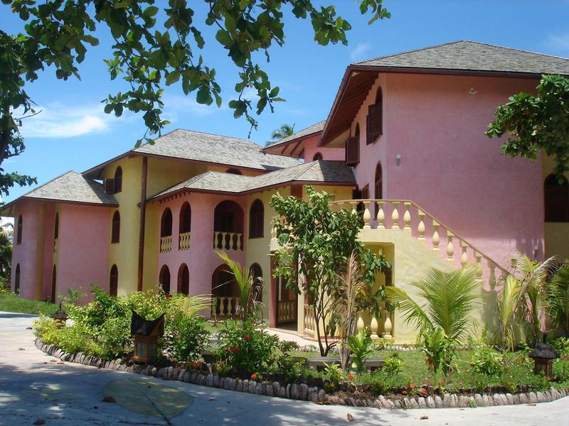 Großes, rosa-gelbes Gebäude mit mehreren Stockwerken, umgeben von grünen Pflanzen und blauem Himmel.