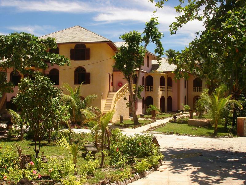 Großes zweistöckiges Gebäude in beiger Farbe, umgeben von Grün und Pflasterweg bei blauem Himmel.