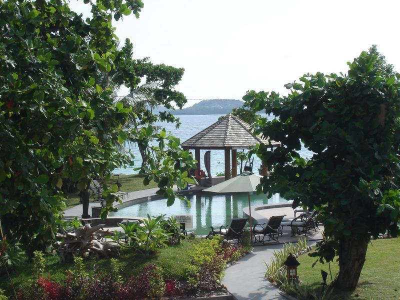 Grüner Garten mit Bäumen und Pavillon am Wasser, dahinter Hügel und heller Himmel.