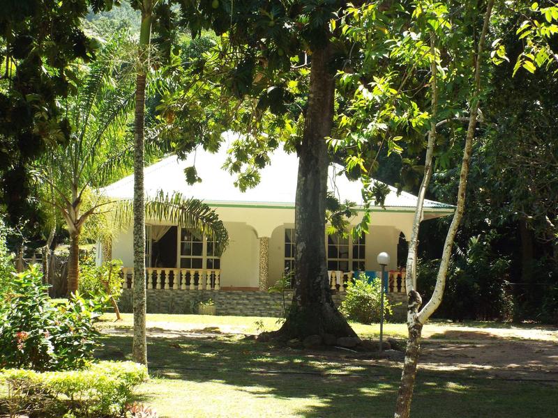 Haus mit heller Fassade, Vordach und Balkon, umgeben von hohen Bäumen und grüner Vegetation.