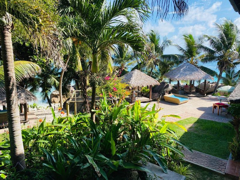 Tropischer Garten mit Palmen und Bungalows nahe klarem Meer unter blauem Himmel.