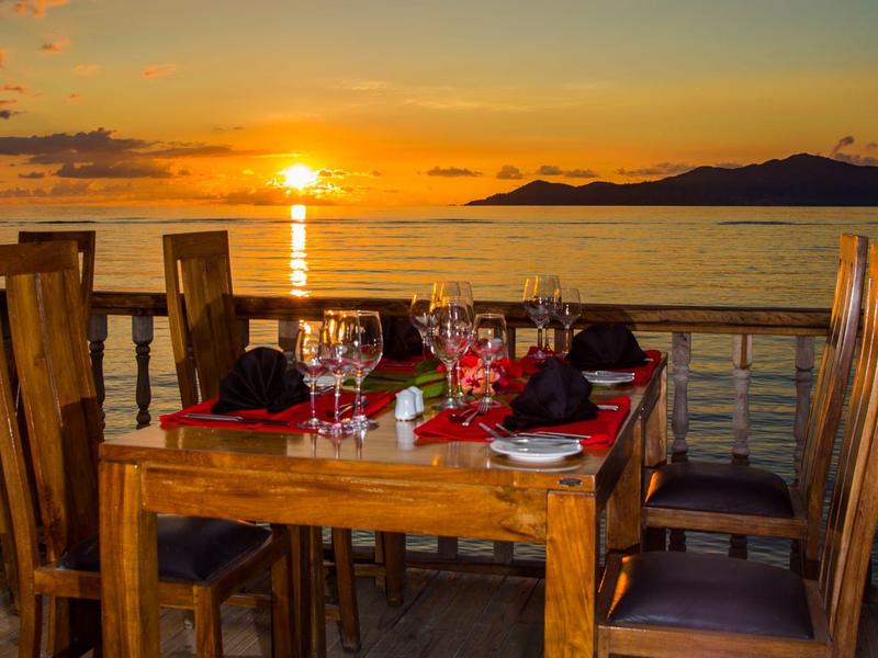 Romantischer Abendessen-Tisch am Meer mit Sonnenuntergang, Holzstühlen und eingedeckten Gläsern.