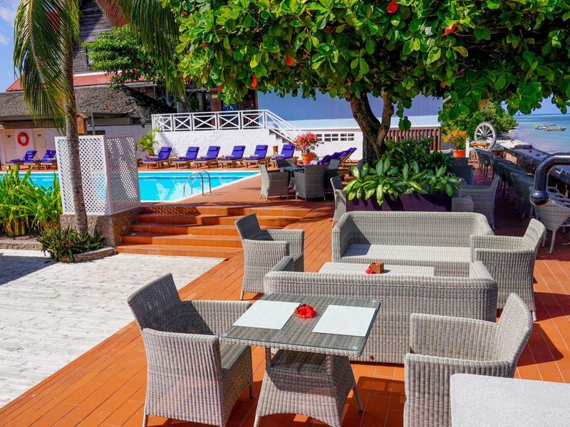 Outdoor-Lounge mit grauen Möbeln, Holzboden, grünen Bäumen und blauem Himmel am Strand.