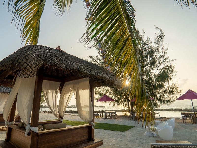 Luxurious pool lounge with curtains under palm trees and sea view at sunset.
